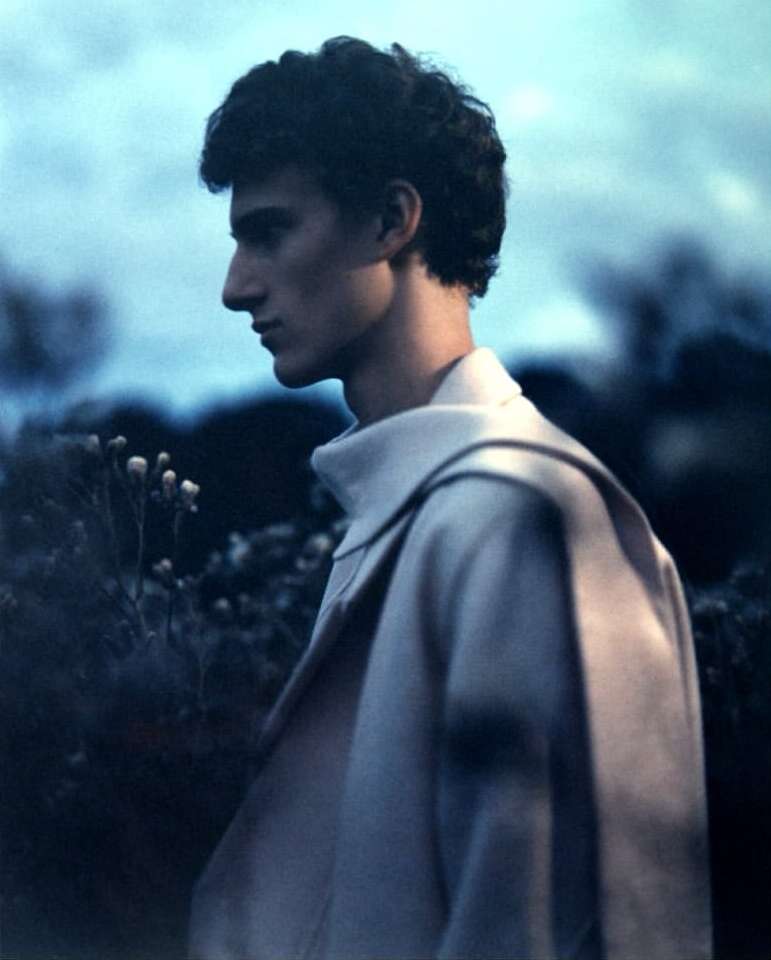 Profile portrait of a young man wearing a light beige coat outdoors against a dark twilight sky by Lluis Camps