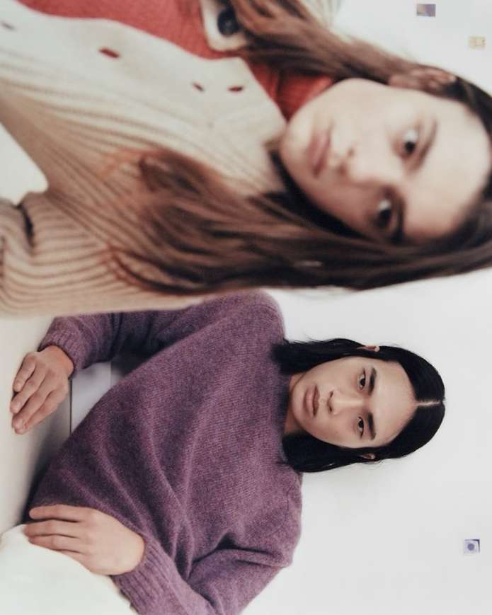 Two people in sweaters lie down facing opposite directions against a white background. One person wears a purple sweater and pants.
