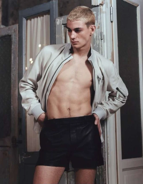 Male fashion model posing with open jacket and black leather shorts next to old door with white curtain in the background.