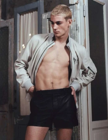 Male fashion model posing with open jacket and black leather shorts next to old door with white curtain in the background.