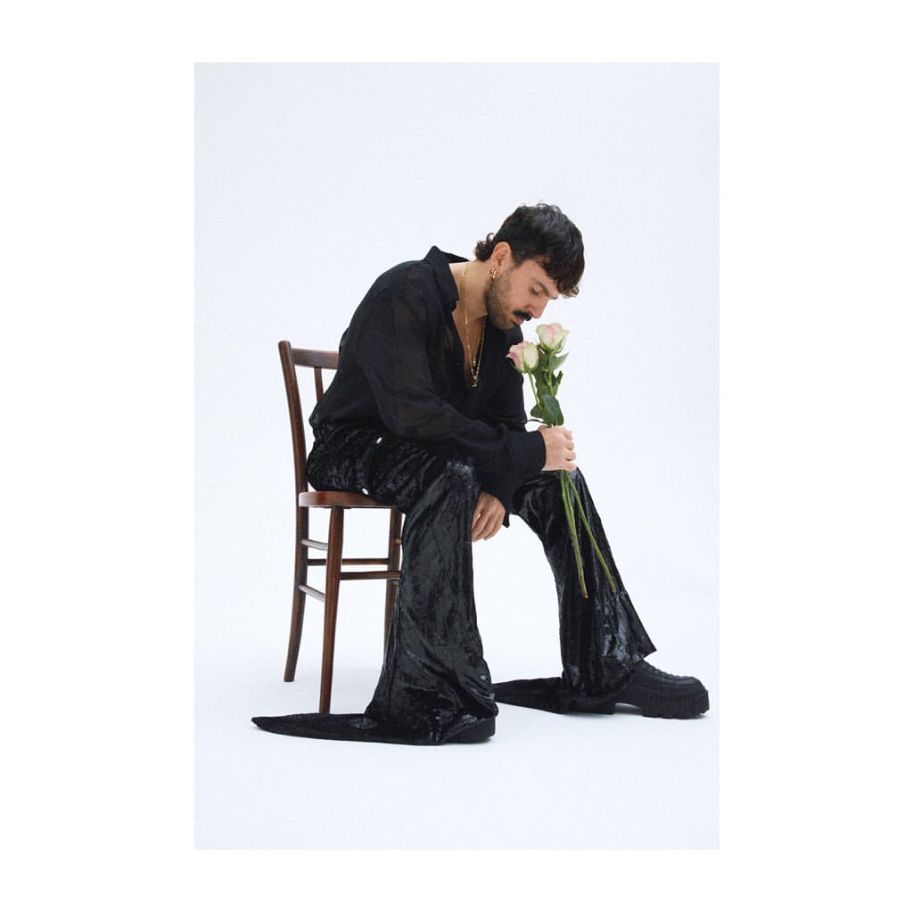 Stylish man in black attire sits on a wooden chair, holding roses, against a clean white background in a portrait composition.