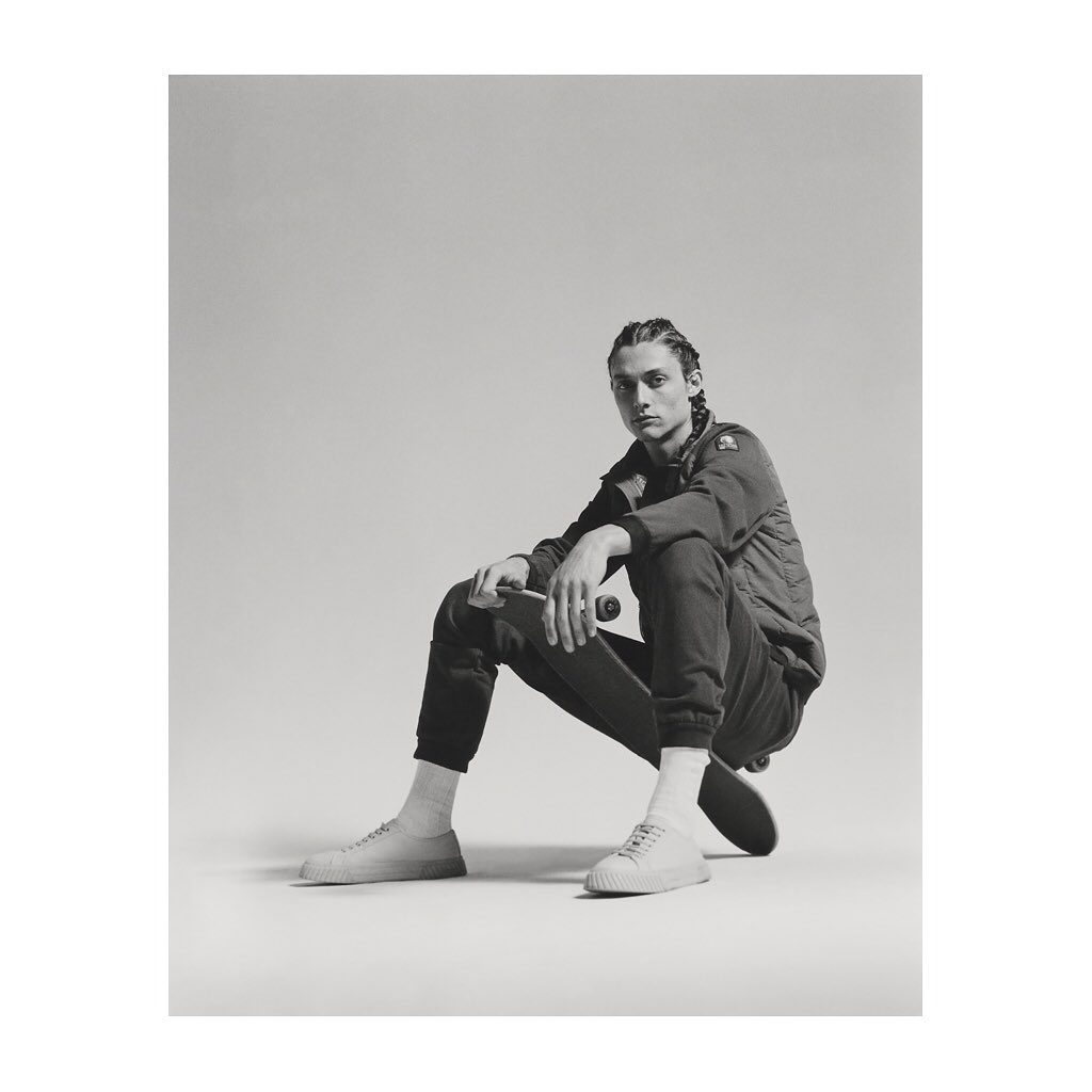 Monochrome shot of a young adult male sitting on a skateboard wearing a jacket, sweatpants, socks, and shoes in a photo studio.