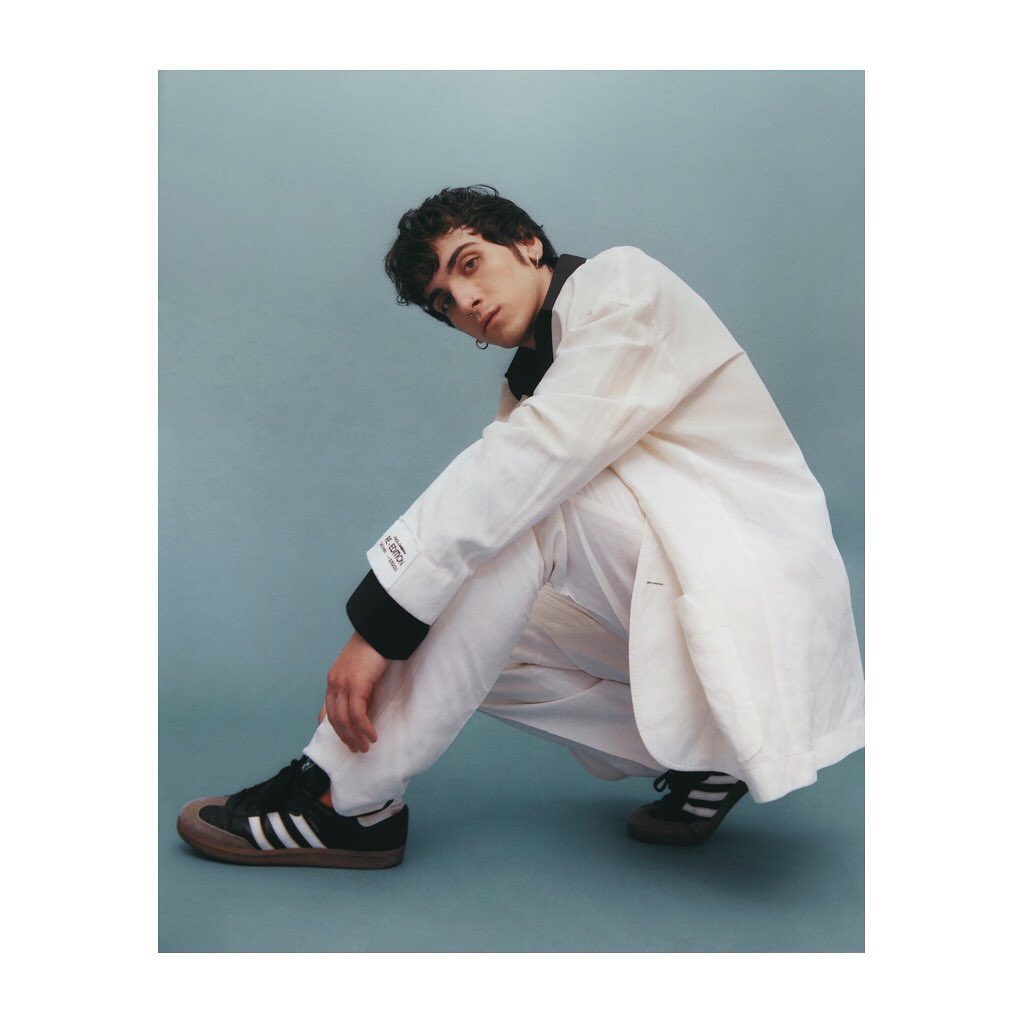 Young adult male in a crouched pose, wearing a white jacket and pants with black and white Adidas sneakers, against a blue background.