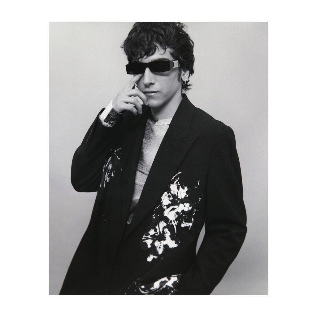 Monochrome portrait of a young adult man wearing a patterned blazer and sunglasses, posing indoors with a neutral background.