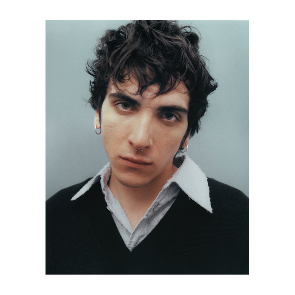 Portrait of a young adult male with curly dark hair, wearing a black sweater over a collared shirt, set against a pale blue background.