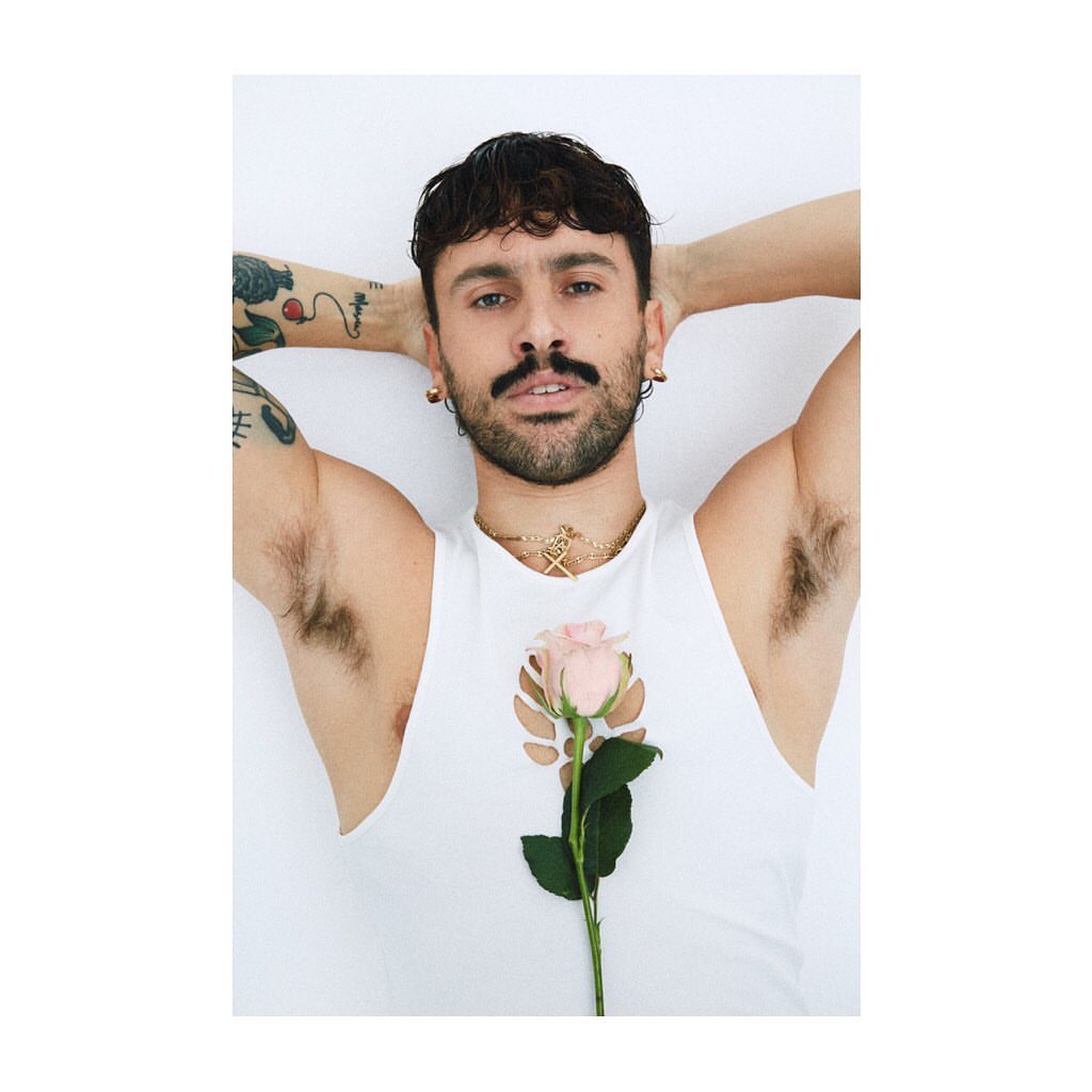 Adult male lying on a white surface with a pink rose. He has dark hair, a mustache, a gold necklace, and tattoos on his arm.