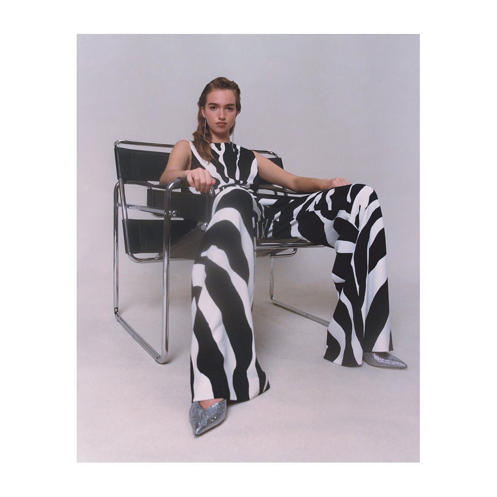 A young woman is sitting in a chrome chair wearing a black and white zebra-striped outfit with silver high heels in a studio shot.