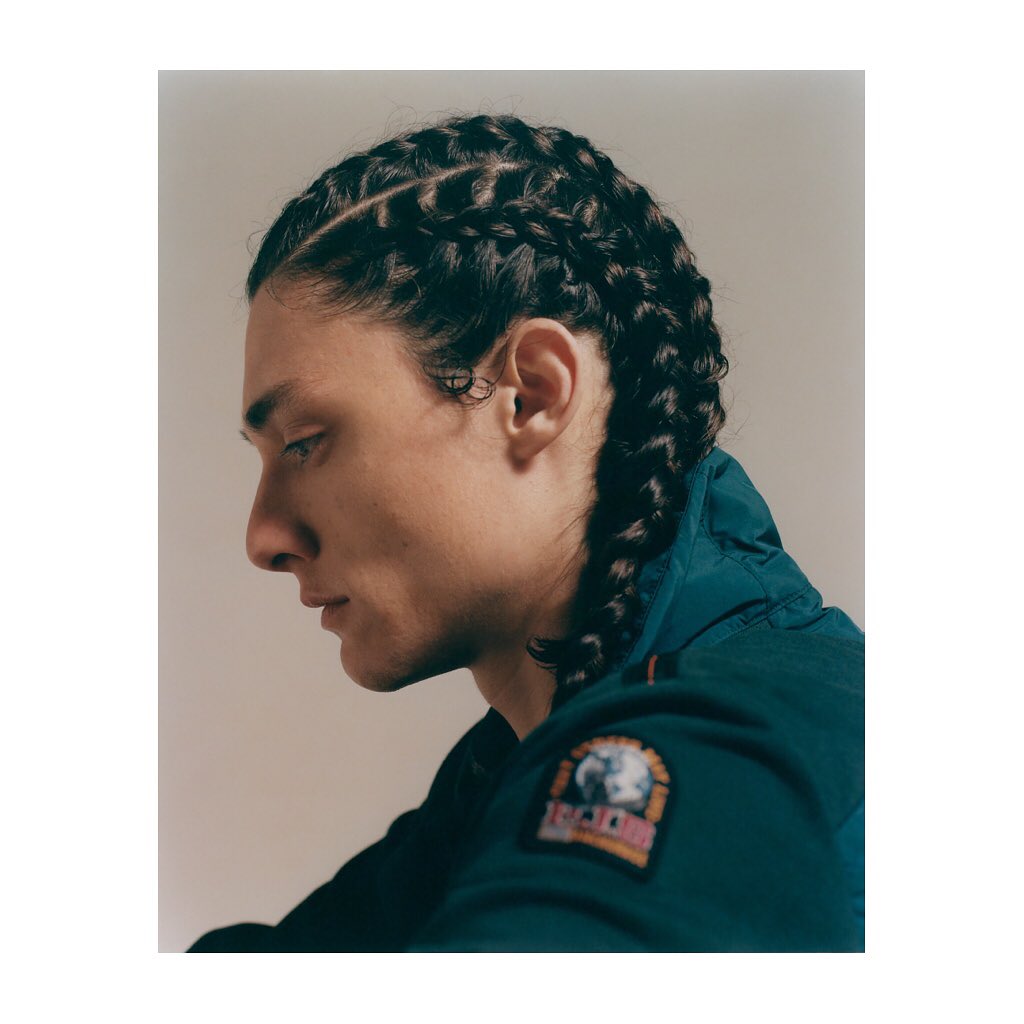 Portrait of a handsome adult male model in a side profile wearing a dark teal jacket with braids against a neutral background.