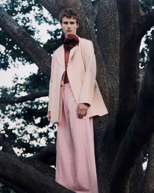 Man in a peach suit stands in a tree. He is wearing a red shirt with a floral piece at the neck, and the background is blurred.