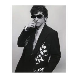 Man wearing dark sunglasses and a patterned black blazer against a light background, photographed in Milan by Lluis Camps.