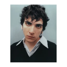 Portrait of a man with dark curly hair wearing a black V-neck sweater and white collared shirt against a grey background.