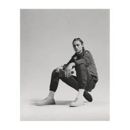 Young man with braided hair sitting on a skateboard wearing a dark jacket, trousers, and white socks against a grey background.