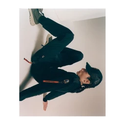 Model wearing dark cap, black long-sleeve top, and matching trousers with an orange tag detail against a plain studio background.