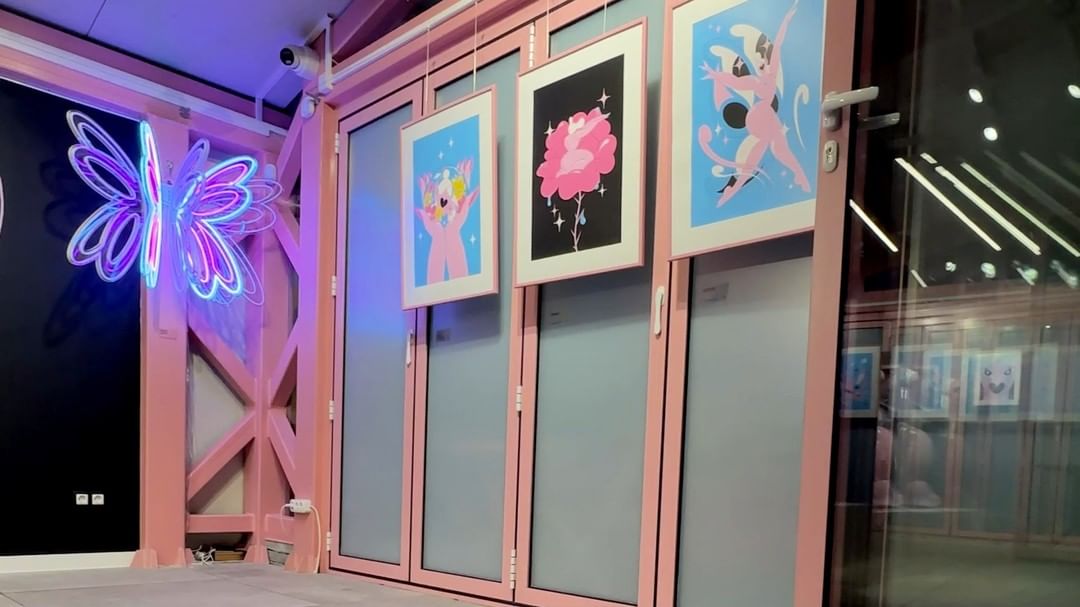 Interior view of a space with artwork displayed on pink-framed walls and a neon butterfly sculpture on the wall, creating an artistic atmosphere.