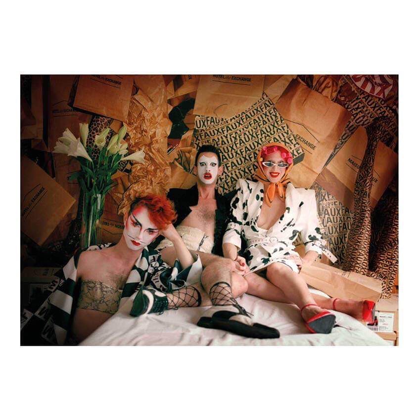 Three drag queens pose in a staged setting with heavy makeup, theatrical costumes, and repurposed shopping bags in the background.