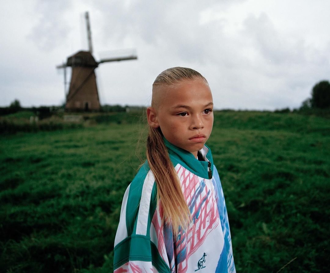 Portrait of a child in a green field, with a windmill in the background under a cloudy sky.