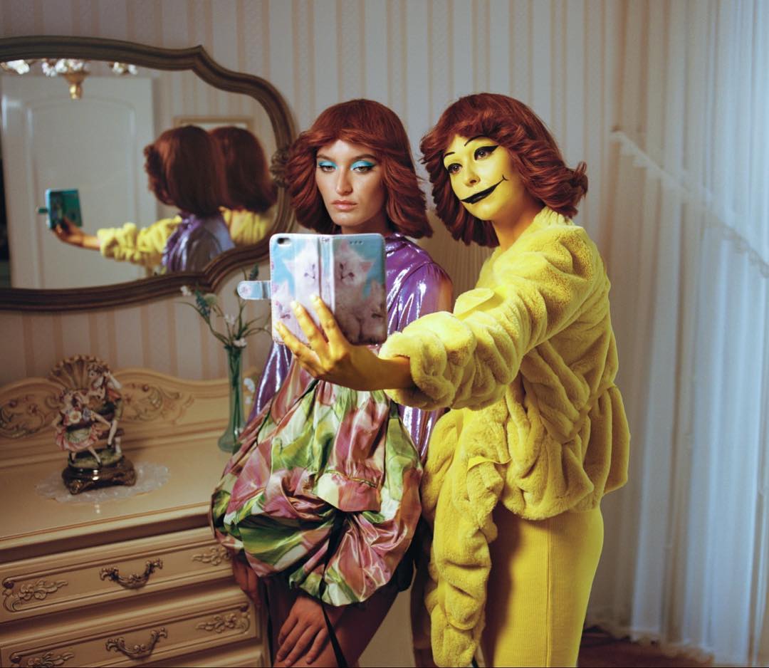 Two women in artistic makeup and colorful fashion take a selfie in an ornate room with a vintage dresser and mirror, vibrant and surreal.