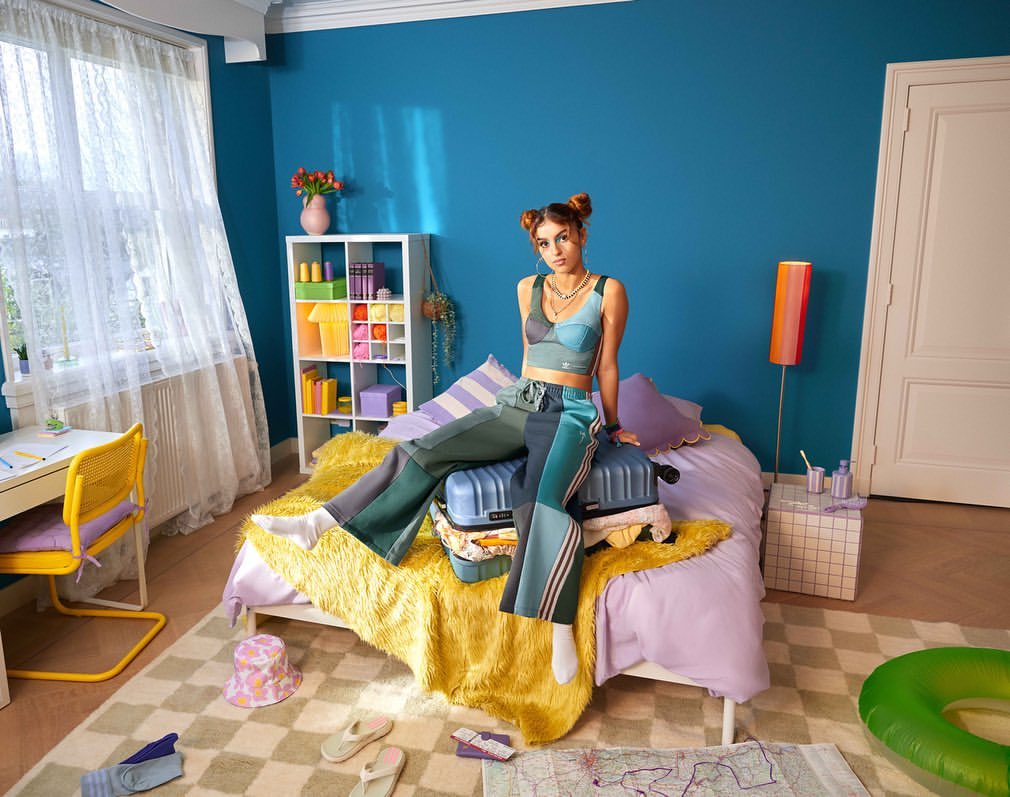Young adult woman with hair buns in teal patchwork outfit sits on suitcases in brightly coloured bedroom with desk and playful decor.
