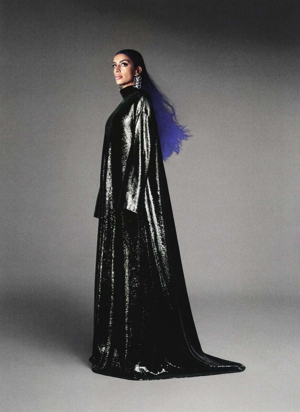 Woman in a black shiny gown poses in front of a grey background with blue highlights in her hair, wearing chandelier earrings.
