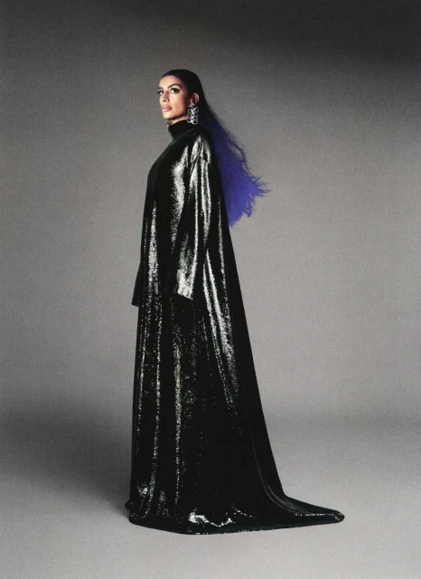 Woman in a black shiny gown poses in front of a grey background with blue highlights in her hair, wearing chandelier earrings.