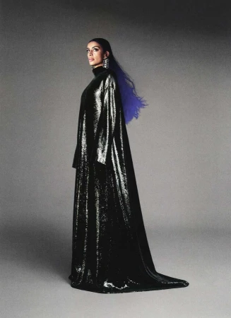 Woman in a black shiny gown poses in front of a grey background with blue highlights in her hair, wearing chandelier earrings.