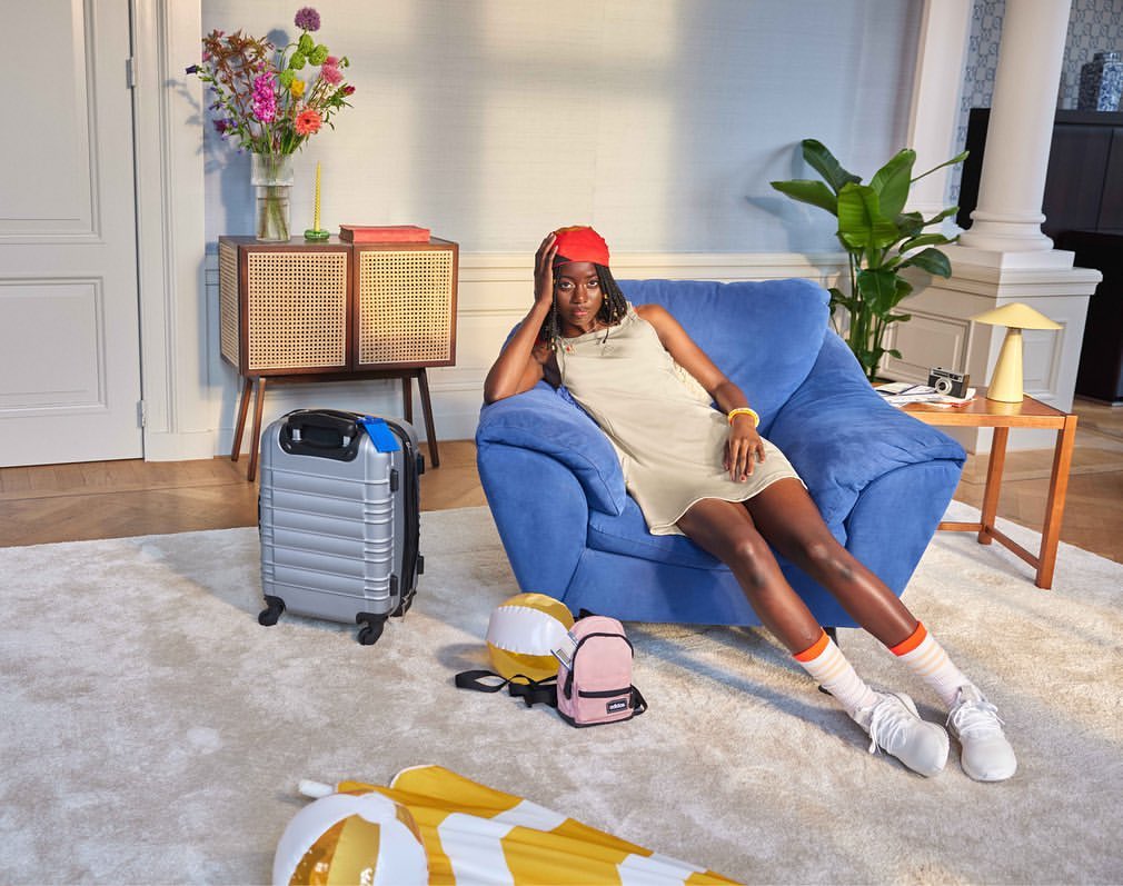 Stylish woman in a dress poses in a chair next to a suitcase, complemented by colorful props, creating a vibrant indoor scene.
