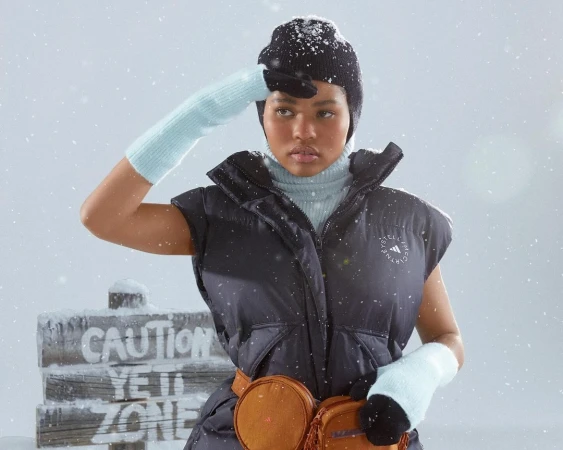 A young woman in winter apparel shields her eyes, standing outside in a snowy setting with a 'Caution Yeti Zone' sign.