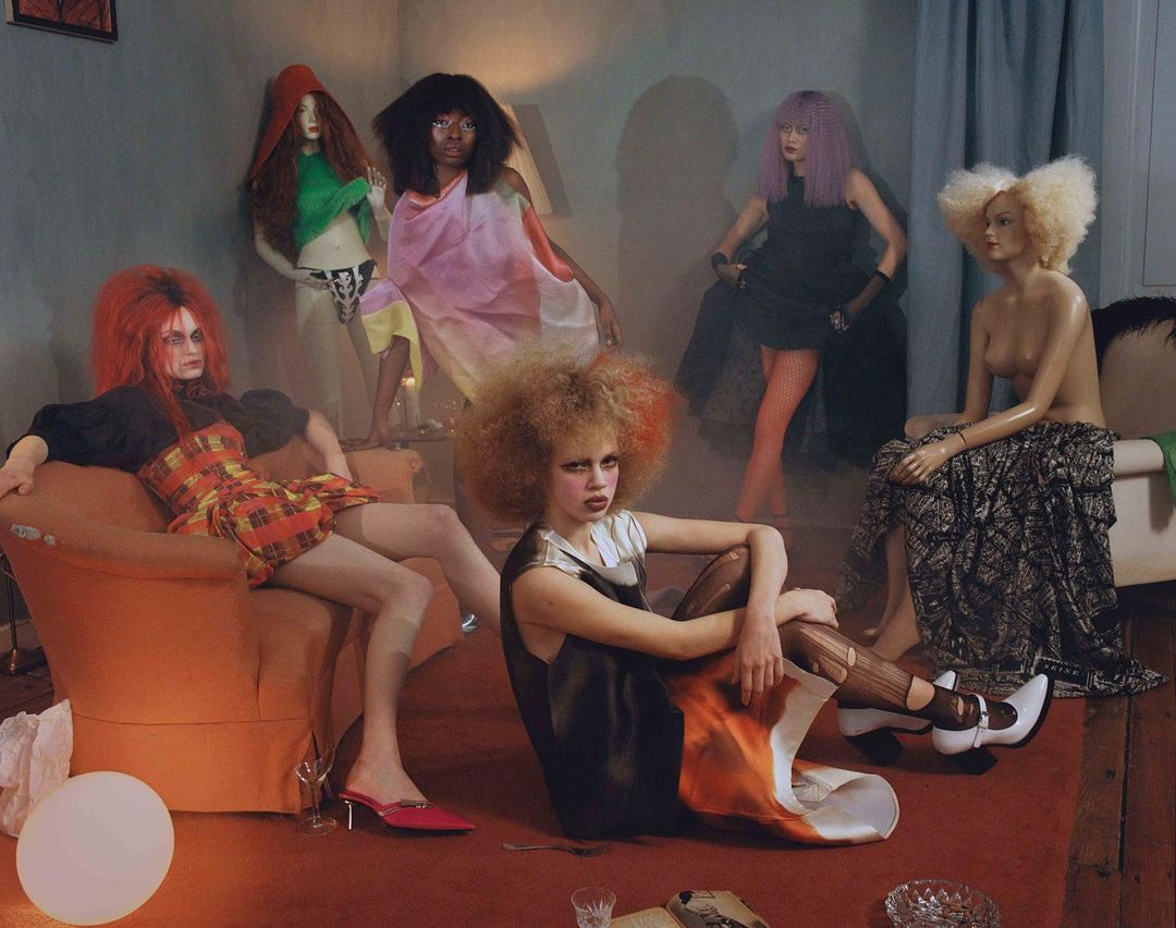 Several fashion models wearing avant-garde colorful outfits and dramatic makeup gather in an indoor scene with a muted color palette.