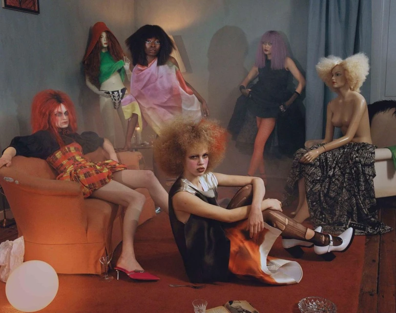 Several fashion models wearing avant-garde colorful outfits and dramatic makeup gather in an indoor scene with a muted color palette.