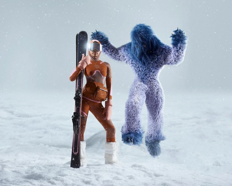 A woman in a ski suit stands with skis next to a blue, furry Bigfoot creature against a snowy background, studio lighting.