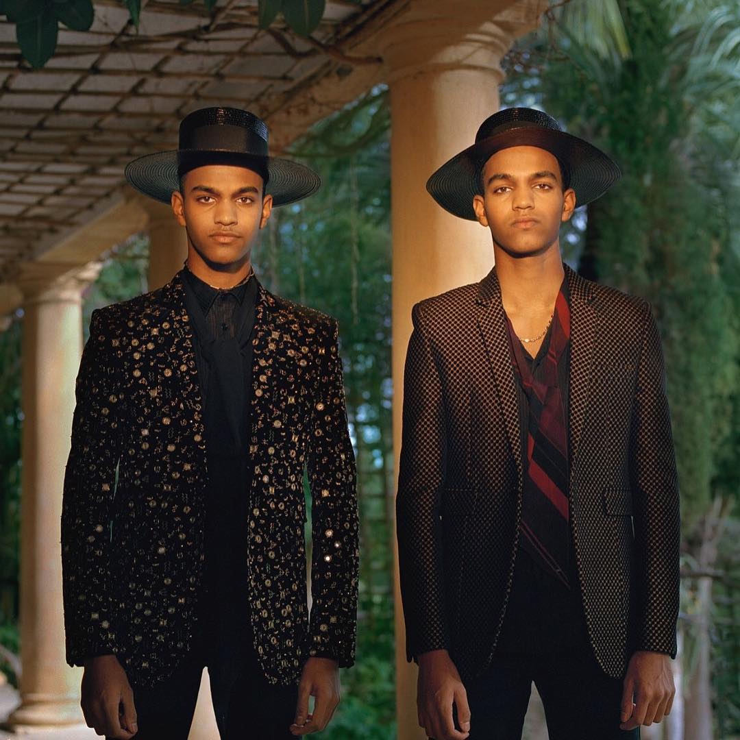 Two adult males in patterned blazers and hats stand in front of an outdoor colonnade with a garden in the background, portrait orientation.