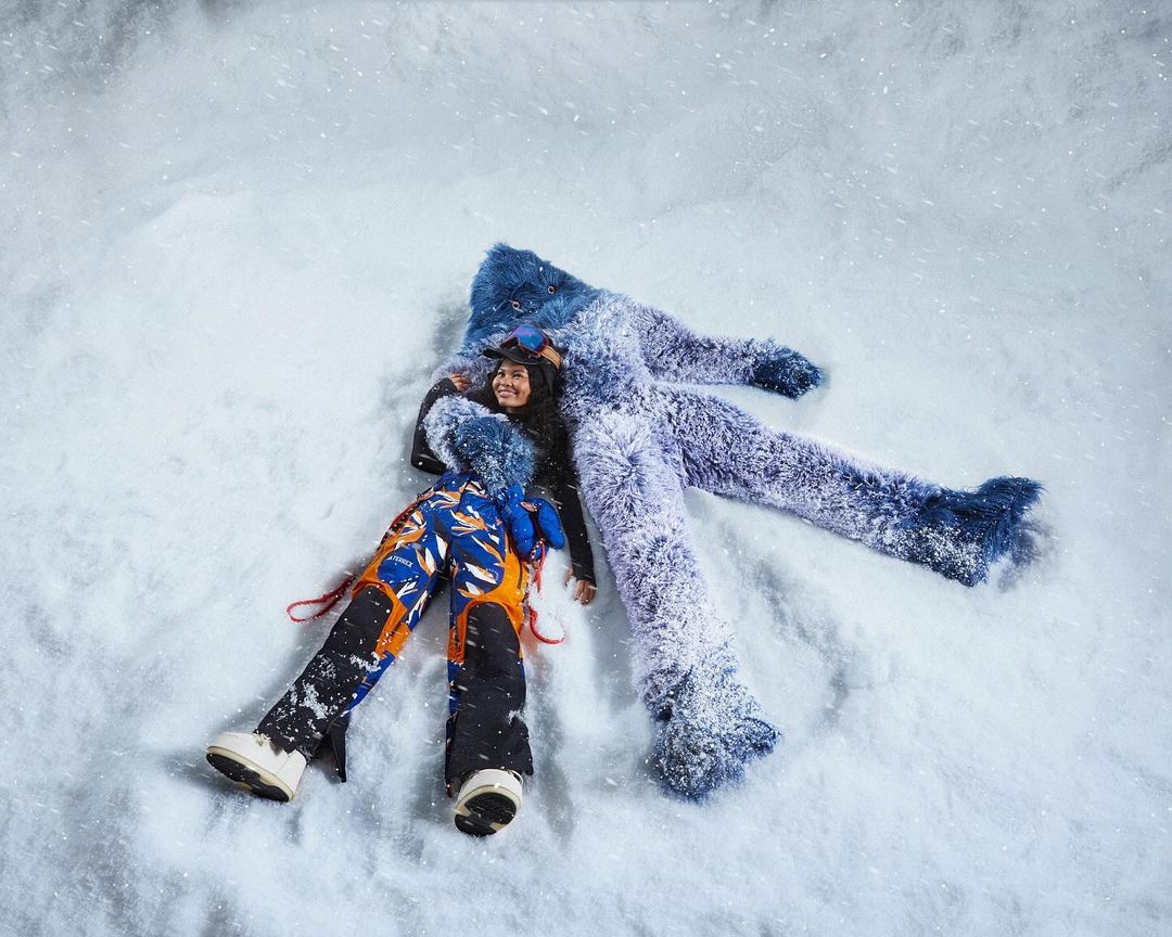 Overhead shot of woman lying in snow making snow angel with fur-lined pants, smiling, wearing colorful ski pants, white snow.