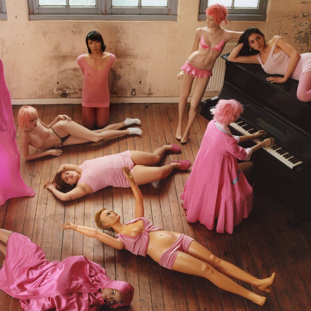 Nine women are posed in pink clothing on a wooden floor with windows in the background, one playing a piano.