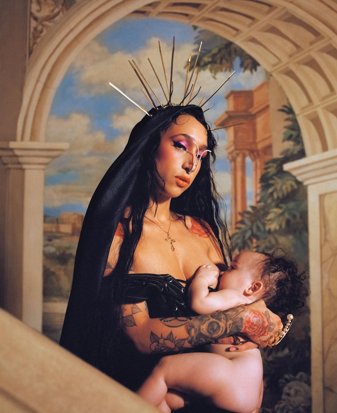 Woman with tattoos wearing a black head covering and holding a baby in front of a painted mural background, portrait orientation.