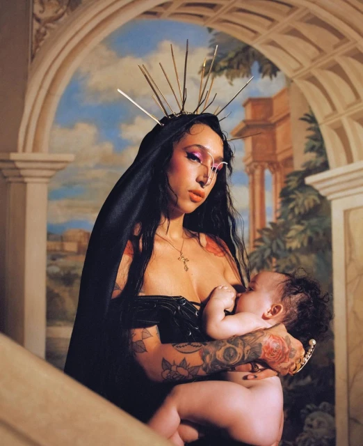 Woman with tattoos wearing a black head covering and holding a baby in front of a painted mural background, portrait orientation.