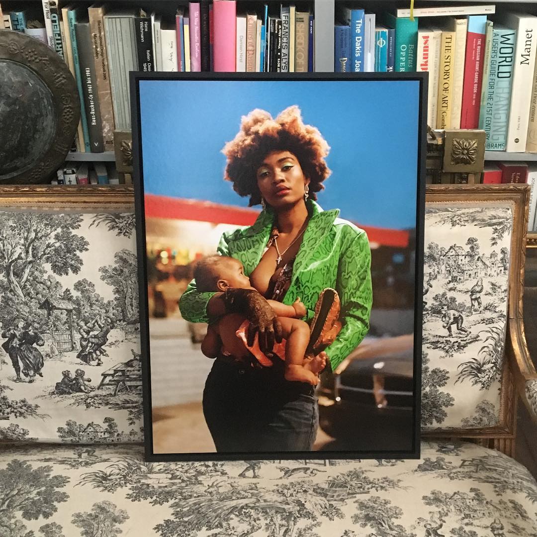 Framed photo of an African-American woman breastfeeding her baby outdoors with a gas station in the background, displayed on a toile-patterned sofa.