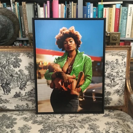 Framed photo of an African-American woman breastfeeding her baby outdoors with a gas station in the background, displayed on a toile-patterned sofa.