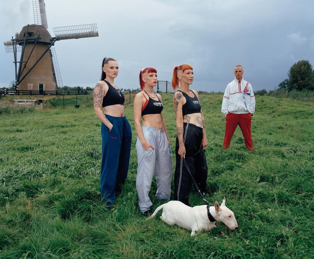 Three women pose in athletic wear with a white bull terrier in a grassy field with a windmill and man in the background.