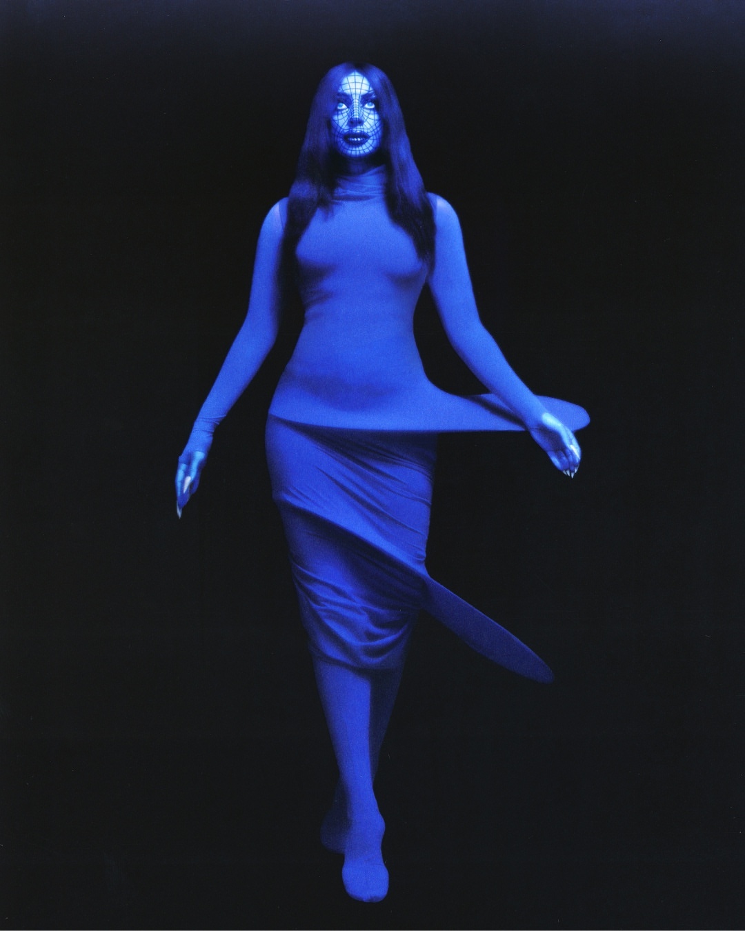 A woman with long dark hair stands in a blue dress against a dark backdrop, creating a striking visual contrast in a fashion forward image.
