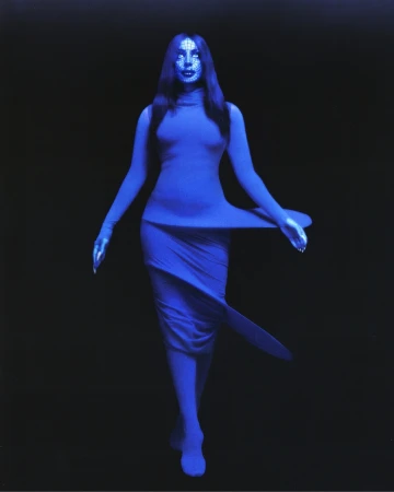 A woman with long dark hair stands in a blue dress against a dark backdrop, creating a striking visual contrast in a fashion forward image.