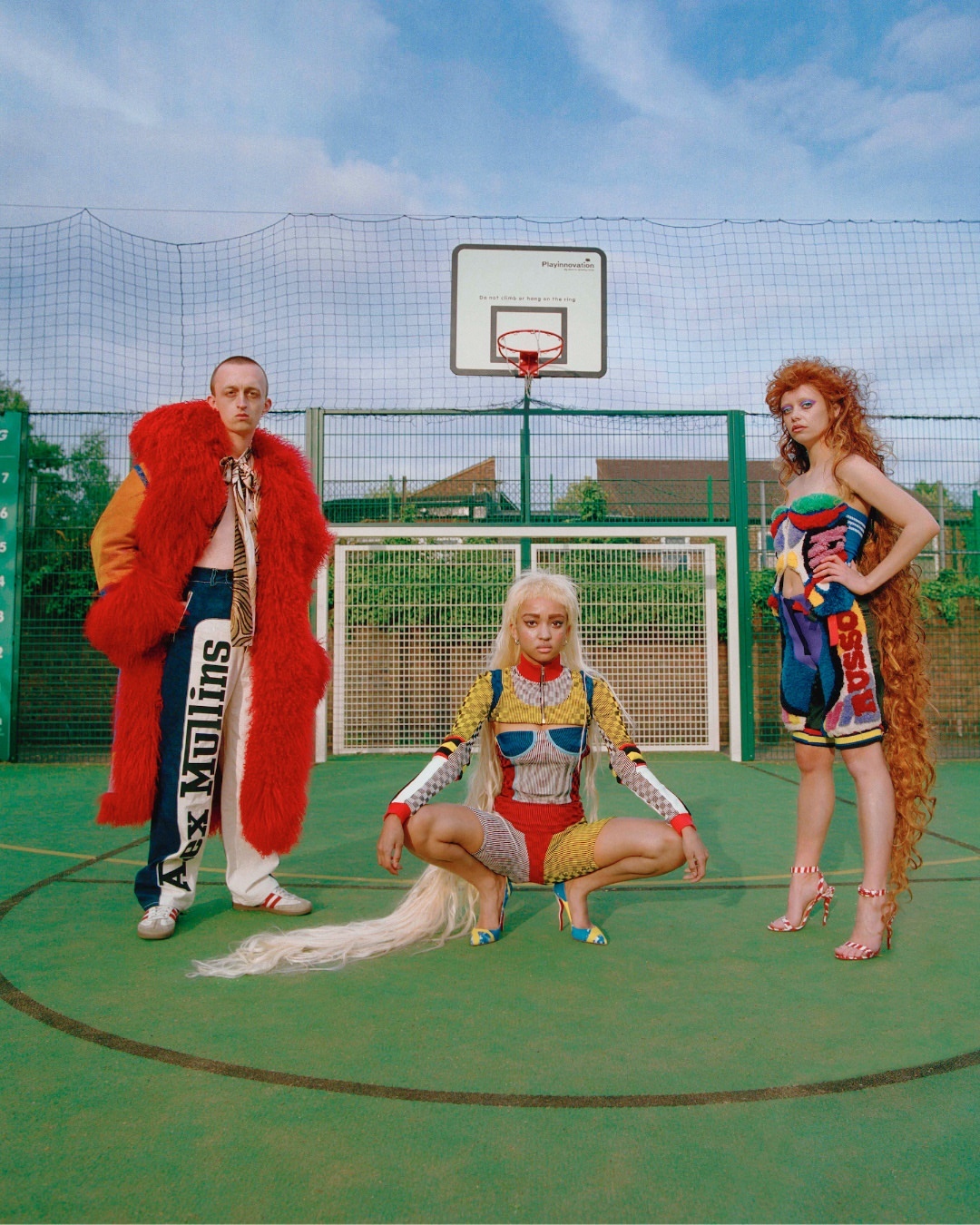 Three fashionable people on a green court: one in a red coat, another crouching with blonde hair, the third with red hair.  