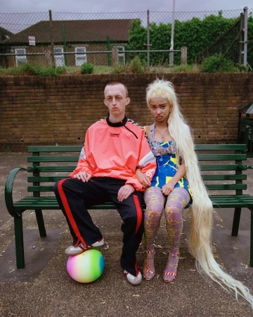 Two people seated side-by-side on a green park bench against a brick wall and chain-link fence under natural daylight.