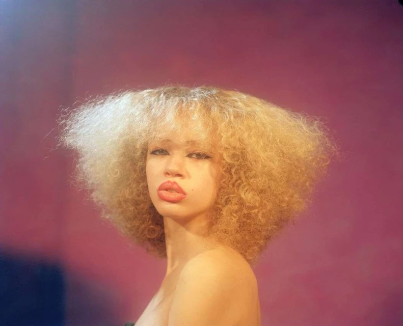 Portrait of a blonde woman with curly hair against a pink backdrop, showcasing beauty and fashion in a studio setting.