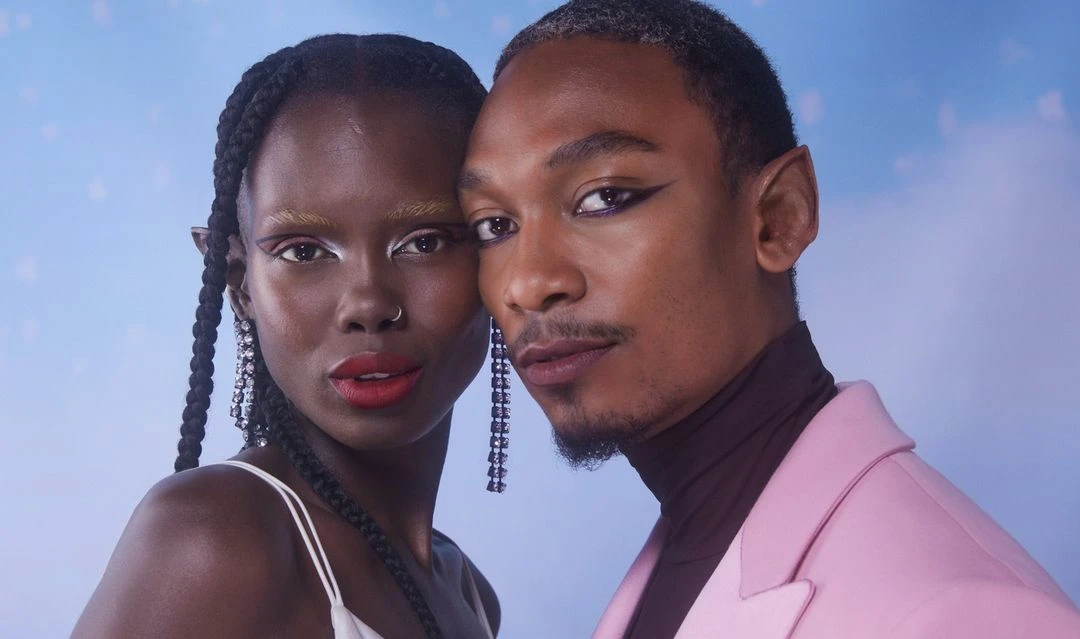 Close-up of a couple with fantasy makeup: woman with braided hair and red lipstick, man in a pink jacket with winged eyeliner.