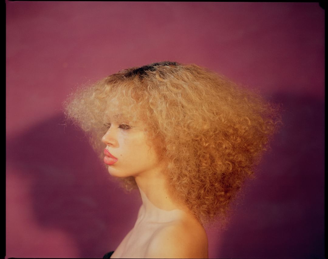 Portrait of a young woman with curly blonde hair against a solid purple background, showcasing makeup and hairstyle in a studio setting.