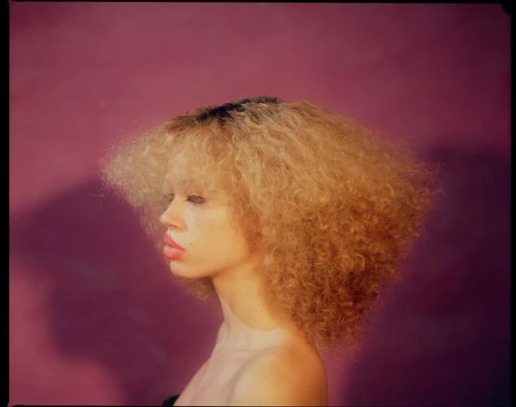 Portrait of a young woman with curly blonde hair against a solid purple background, showcasing makeup and hairstyle in a studio setting.