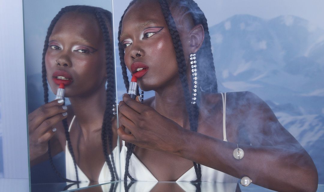 Woman with dark skin and braided hair applies red lipstick in front of a mirror against a cloudy, mountain backdrop with light blue filter.