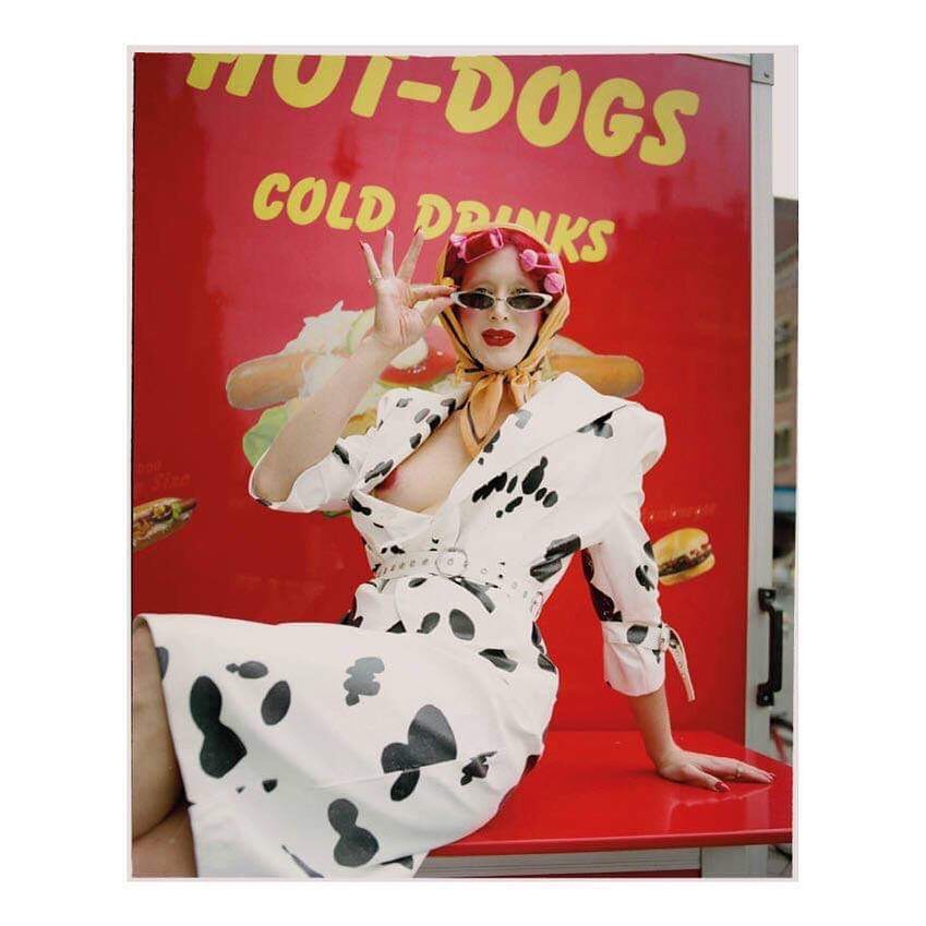 Woman in patterned dress and retro sunglasses sitting in front of a food truck with hot dog and cold drink advertising.