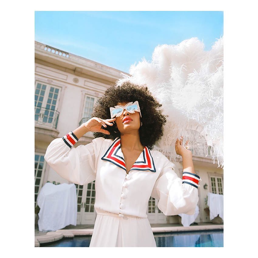 Stylish woman poses in sunglasses with white feather fan against a building and blue sky backdrop; outdoor fashion portrait.