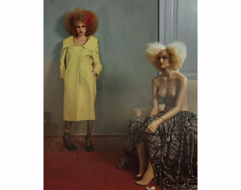 A posed fashion model with an afro hairstyle wears a yellow coat next to a mannequin in an indoor setting with minimal decor.